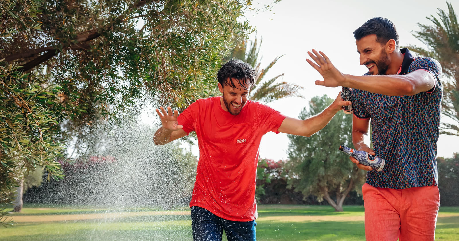 Two men laughing in a garden sprinkler, one in a red A Fish Named Fred tee